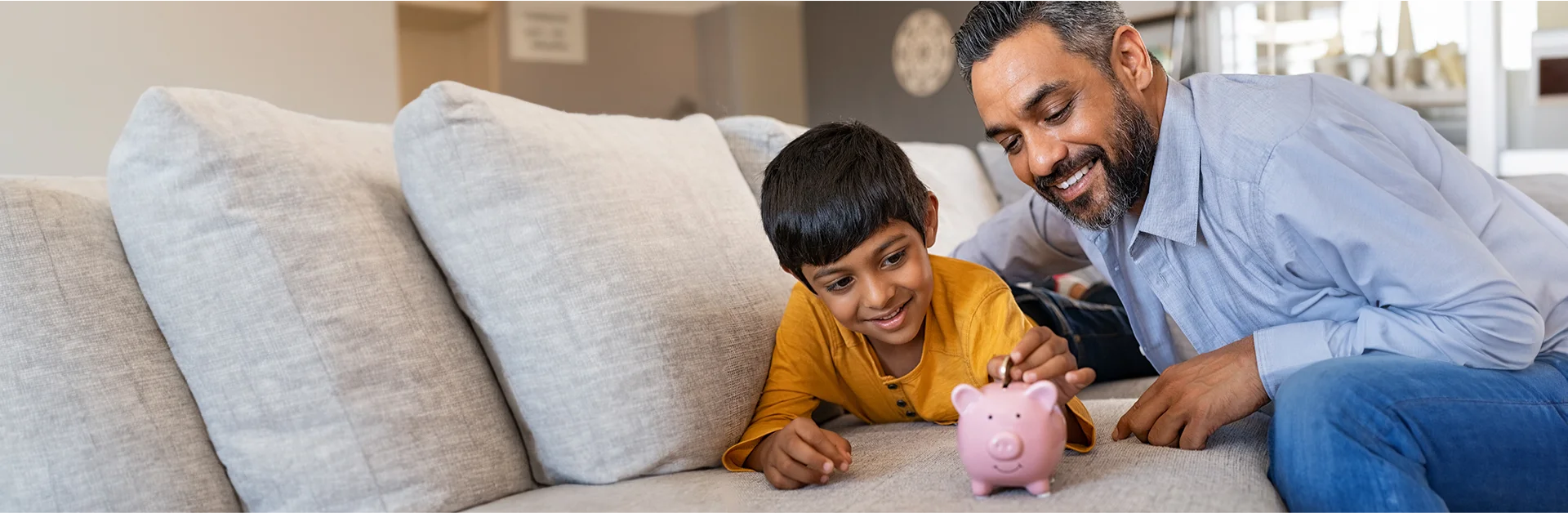 Man and son lying on couch putting coin into piggy bank